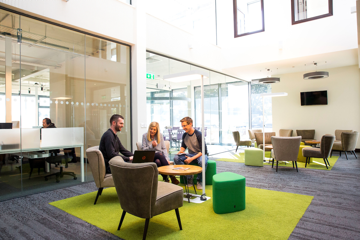 3 people sit around a circular table discussing something on a laptop that sits in front of the 3 of them. They are in the middle of a large, open office space