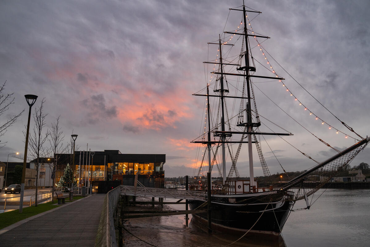 To the centre-right of the image is the Dunbrody Famine Ship, docked on the Barrow River which runs through New Ross town. To the left of the image is the boardwalk that leads into the Dunbrody Famine Ship Experience tourist centre. The sky is cloudy with a pink sunset peeking out from the clouds