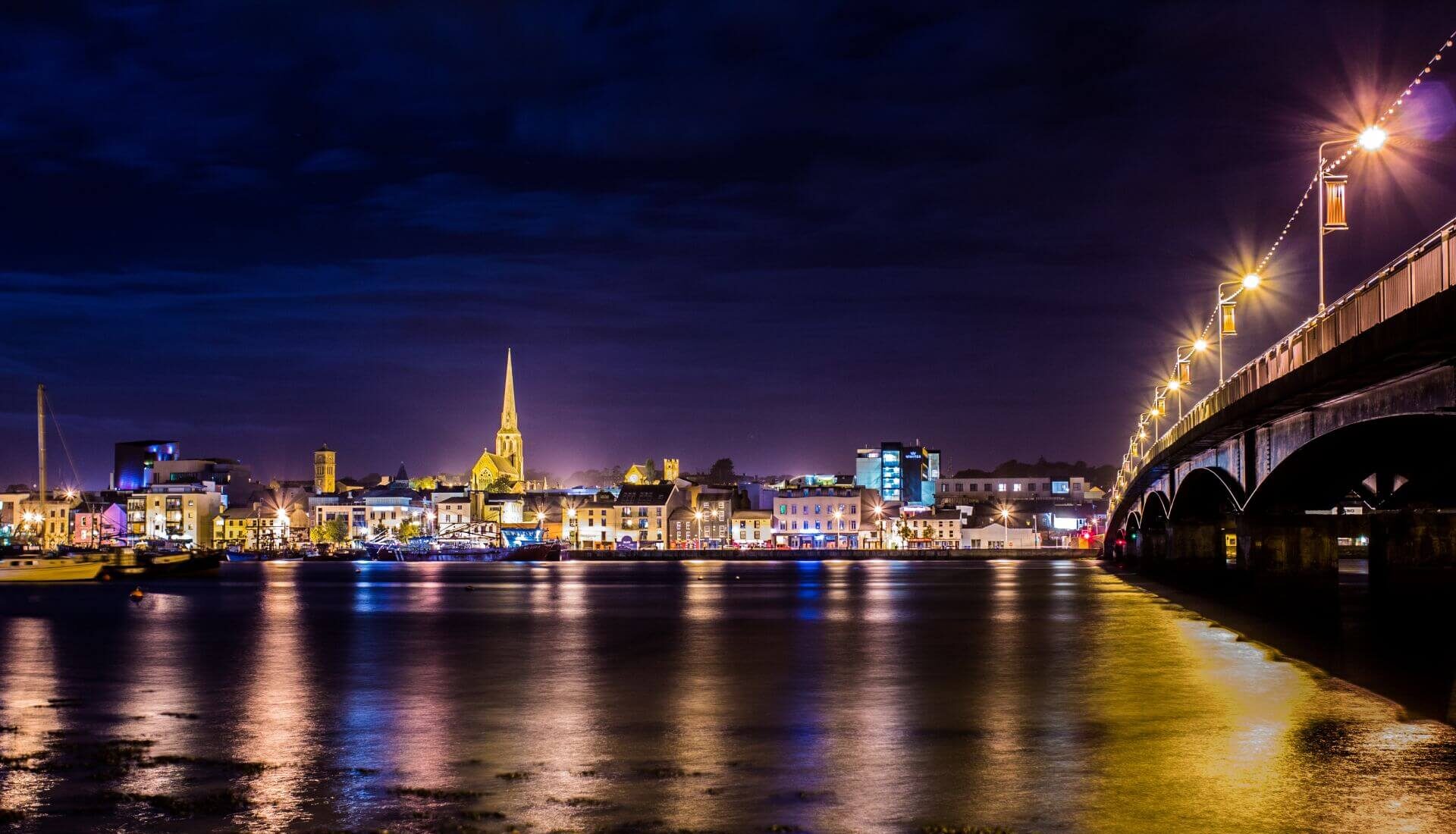 Night time photo of Wexford Town, taken from across the main bridge looking back into the town. It is illuminated by nightlife and features the town's skyline with a church spire, hotels, bars and boats in the background and the reflection of the lights bouncing off the River Slaney in the foreground. To the right of the photo is the Wexford bridge with bright streetlights beaming down