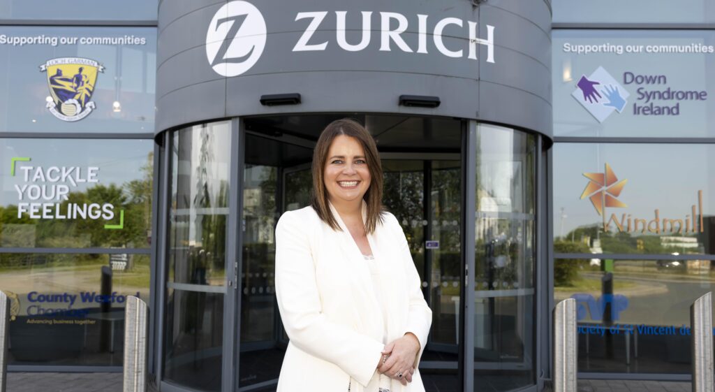 In the middle foreground is Zurich Insurance's Chief Compliance Officer & Head of EEA Oversight at Zurich, Sarah Pierce. She is wearing a white blazer and smiling widely. She stands in front of the modern, rotating door that brings you into the Zurich Wexford office. On the windows are graphics of the businesses and charities that they support which includes: Wexford GAA, Tackle Your Feelings, Down Syndrome Ireland, St Vincent De Paul and Windmill Wexford. They also have a County Wexford Chamber sticker to show their membership