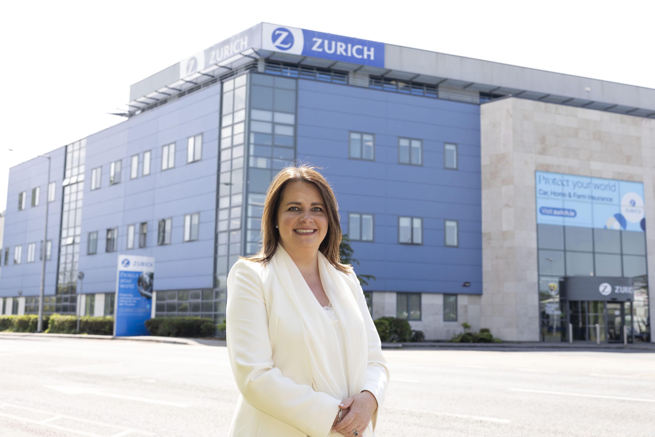 In the middle left foreground is Zurich Insurance's Chief Compliance Officer & Head of EEA Oversight at Zurich, Sarah Pierce. She is wearing a white blazer to contrast the multi-storey blue, Zurich office building behind her.