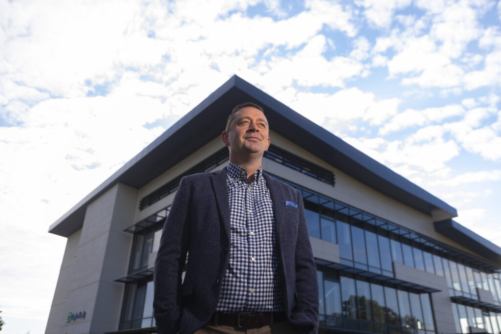 Paul Whelan, Managing Director of Depositary Services for CSC’s Global Financial Solutions stands at the centre of the image in front of the M11 Business Campus in Gorey. He wears a black and white checkered shirt and a navy blazer. He is looking away from the camera