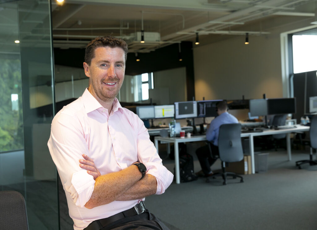 Jim Hughes of Innovate sits at the front left of the image. He is smiling at the camera with his arms folded to the right background is his office space in M11 Business Campus, Gorey. There are multiple desks set up with multiple monitors and a man sits at his desk working