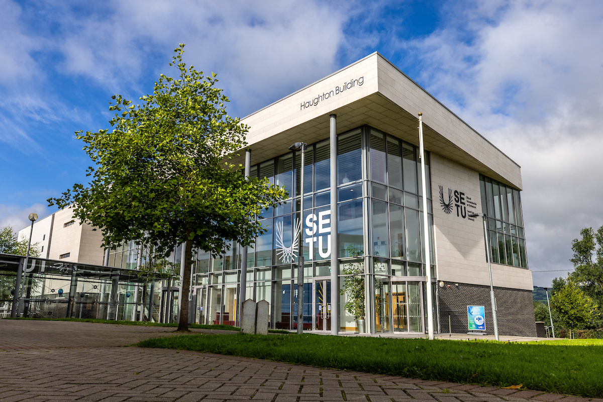 A modern glass front building on Carlow's SETU Campus. The facade says Haughton Building and there are decals of the SETU university logo on the glass-front and stone front of the building.