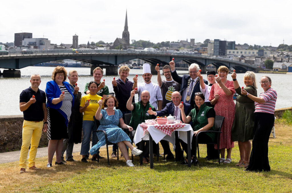 The Wexford Food Producers Network members celebrating the launch of the Wexford Fleadh 2025 on a grassy piece of land in Wexford Town, behind them is the River Slaney,the Wexford bridge and the quayfront of Wexford town