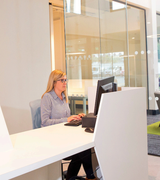 A lady sits at in a bright modern office space with floor to ceiling glass walls to the right of the image. She is looking at a computer monitor