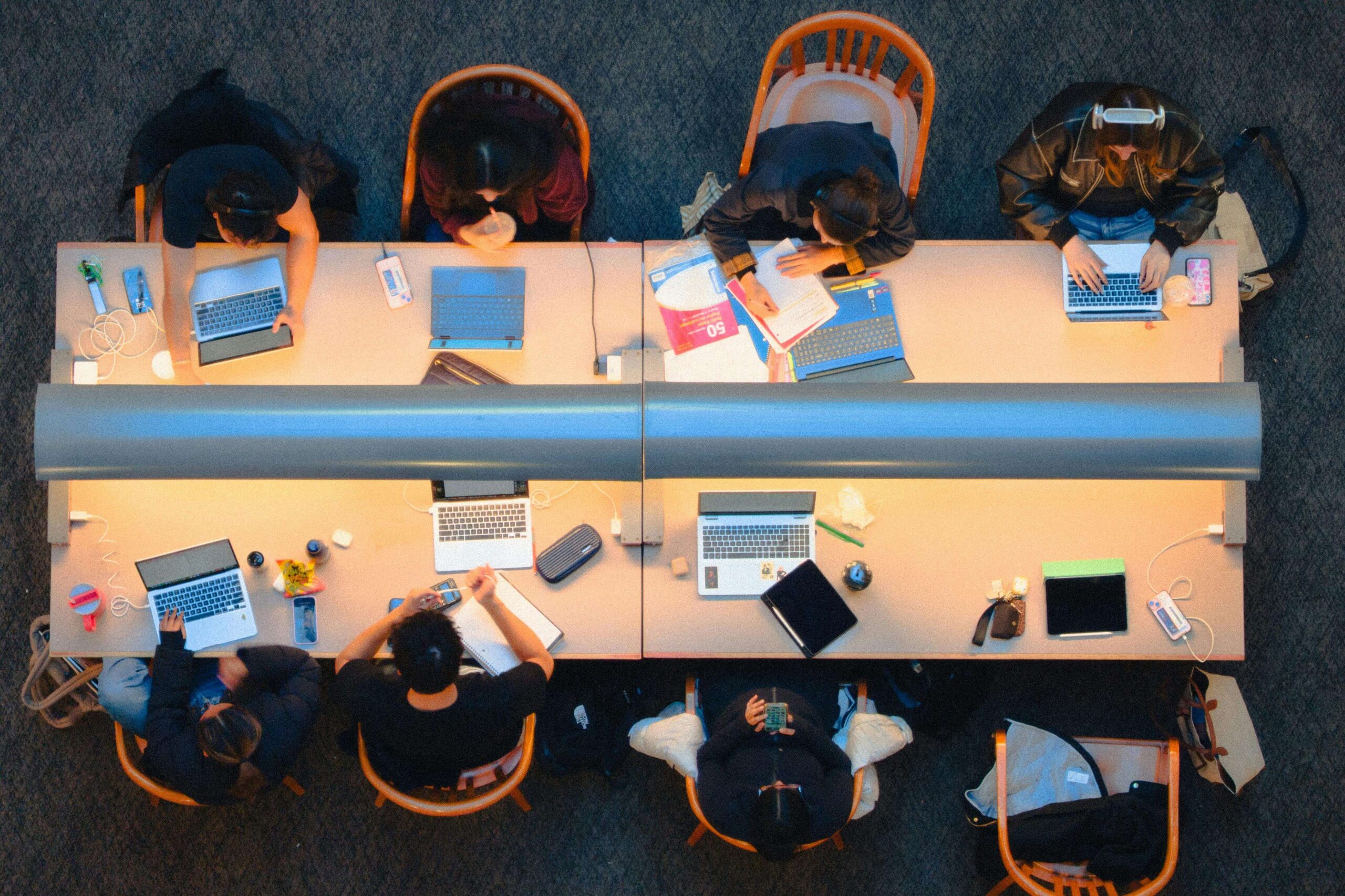 An overhead view of a long library table in a university library, there are 4 chairs either side of the table and a long desk lamp separating both sides. There is an array of laptops and study materials along the desk
