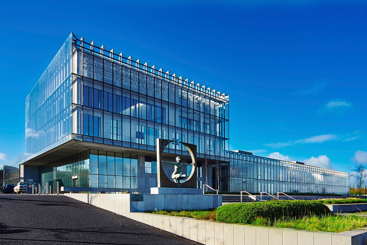 A front view of the Wexford County Council offices in the County Hall. The building is a multi-storey, glass fronted building with exposed metal rods as part of the façade. To the front of the building in the left foreground is a sculpture of a 3d square with a circle cut out in the centre. In the centre is a life-like sculpture of a lady with her arms out, holding both sides of the square. The piece is by Rowan Gillespie and is called 'Solstice'