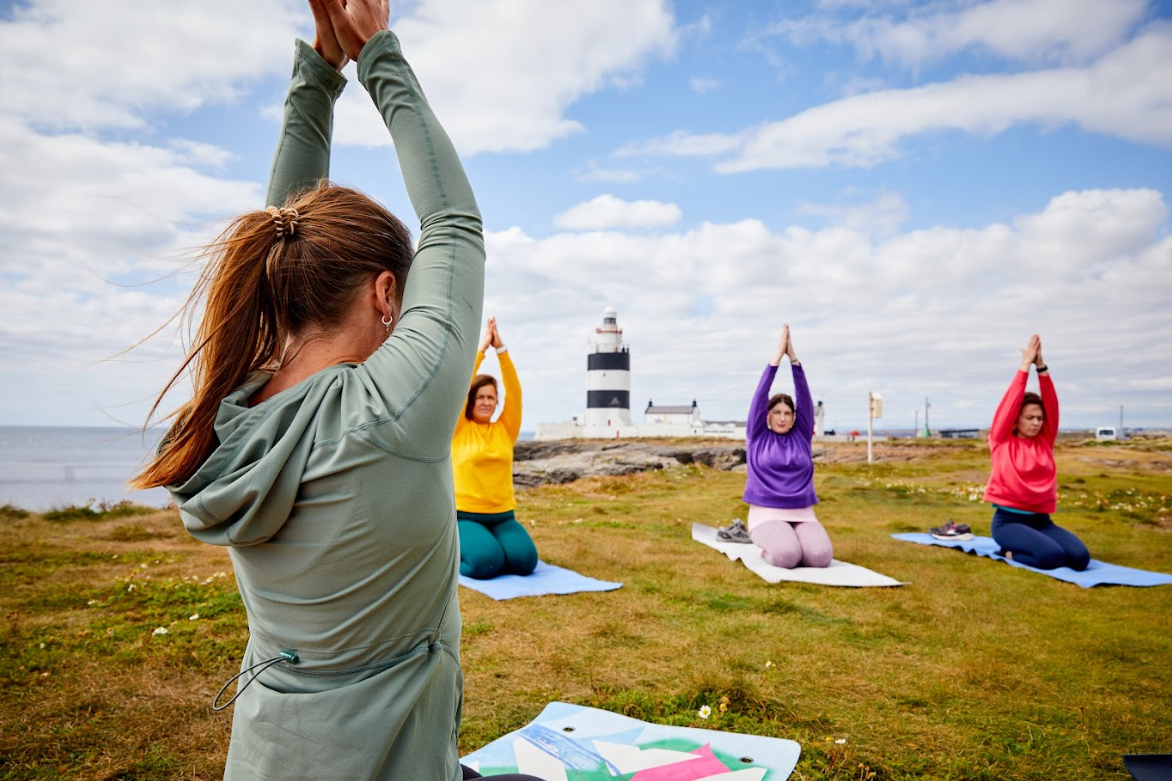 a group of women doing yoga on the Hook Peninsula, in the background is the Hook Lighthouse. They are arranged in a circle, each kneeling on yoga mats and following the instructor on the left of the image. The have their arms over their heads with palms together
