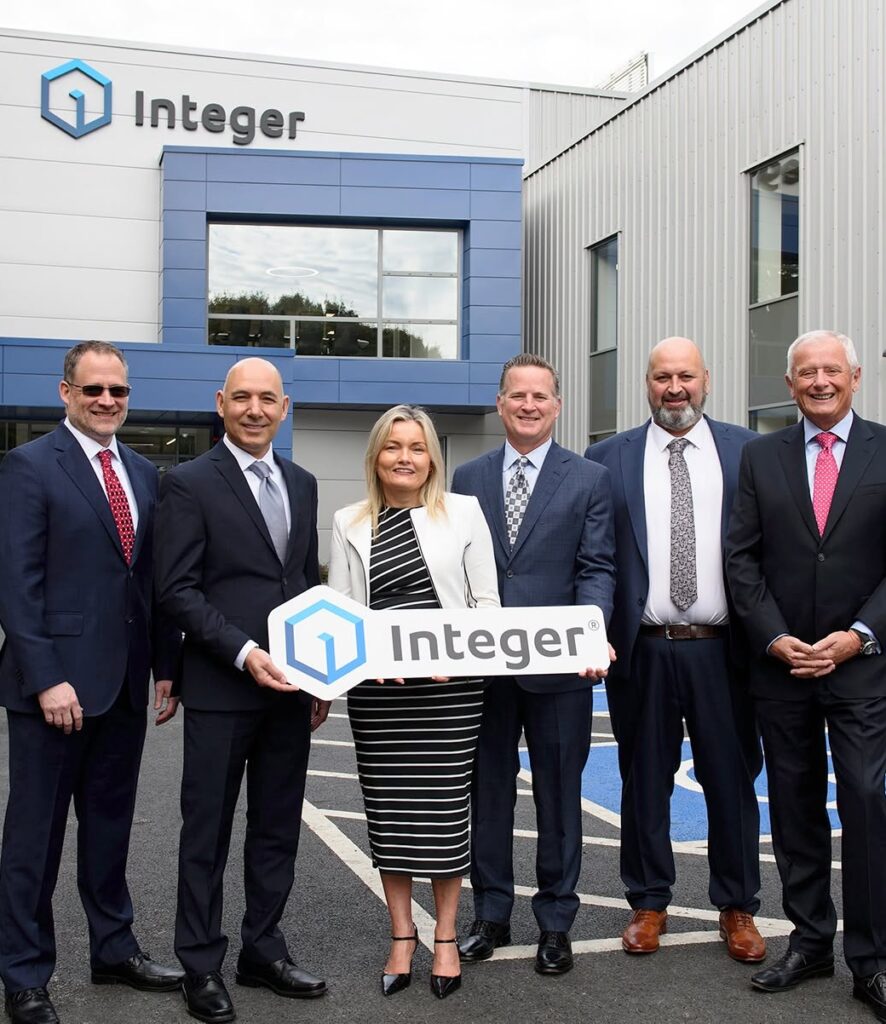 6 people stand in the foreground in front of the new Integer building to celebrate it's opening. There are 5 men and 1 woman, the woman stands in the middle holding a cut out of Integer's logo.