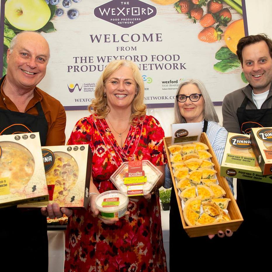 Members of the Wexford Food Producers' network stand in front of a welcome sign at the Wexford Fleadh 2025. They are holding products belonging to Trudie's Kitchen and Zanna Cookhouse