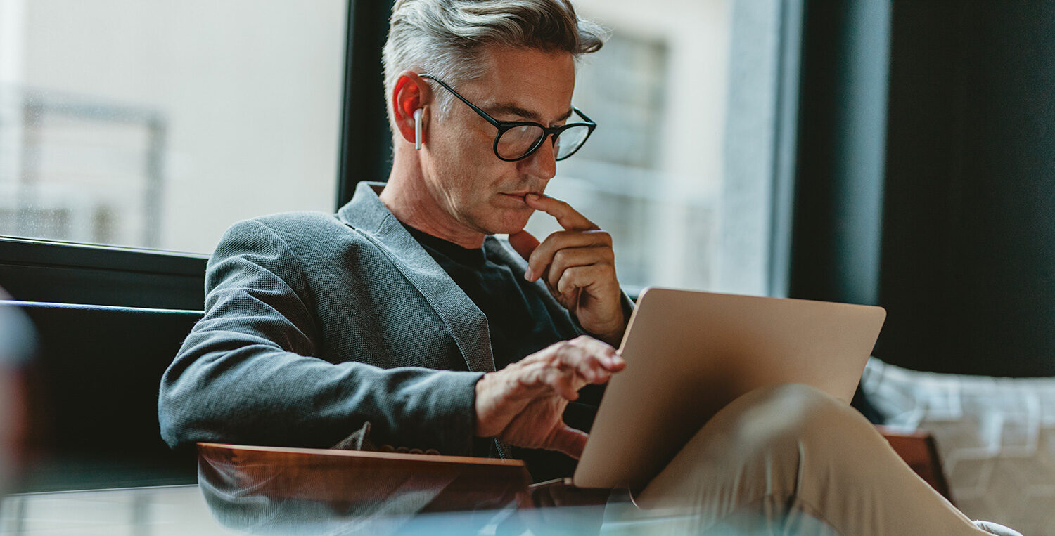 In the centre of the photo, there is a man working on his laptop with earphone in his ears. He holds a finger to his mouth as he is working and behind him there is a large window. He is close to the camera.