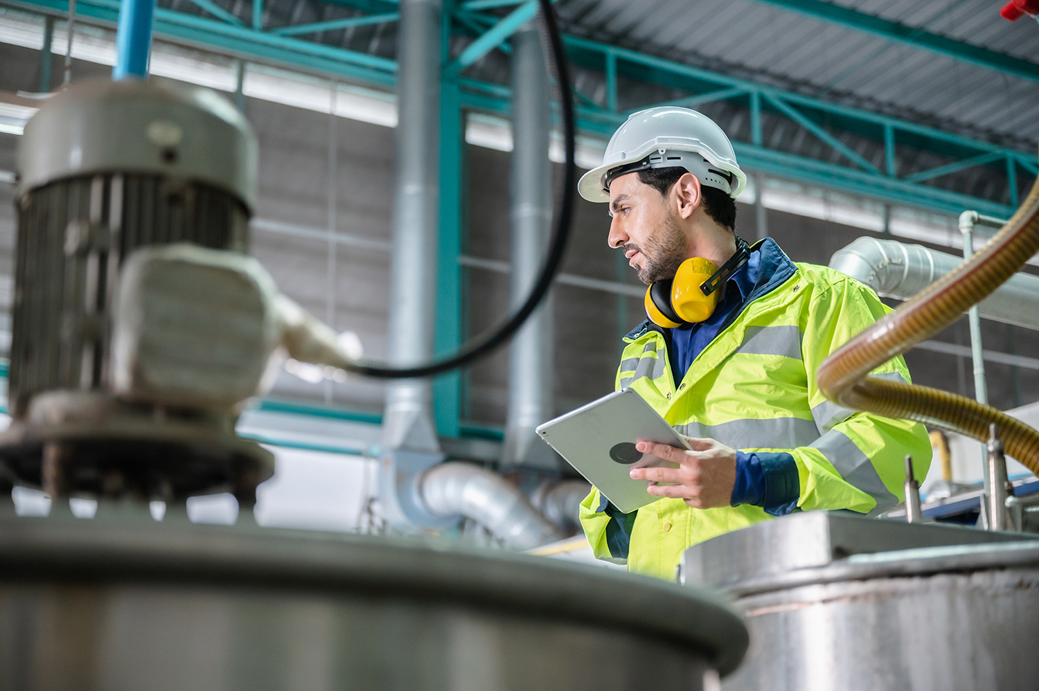 This is an image of a male worker in an industrial facility. In the background there are turquoise blue support beams. In the foreground there are industrial machines to the left. The man is wearing a hi-vis coat, a helmet and has ear defender headphones around his neck. He is holding a tablet and looking to the left of the photo, examining the equipment