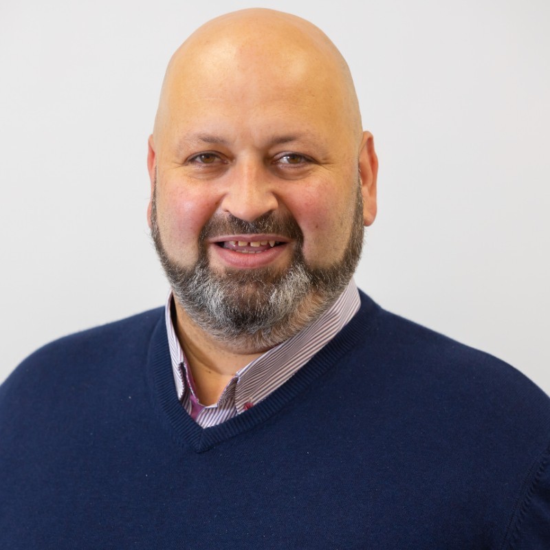 John Mehigan of Integer smiles at the camera, he is wearing a pink striped shirt and navy jumper and is set against a grey background