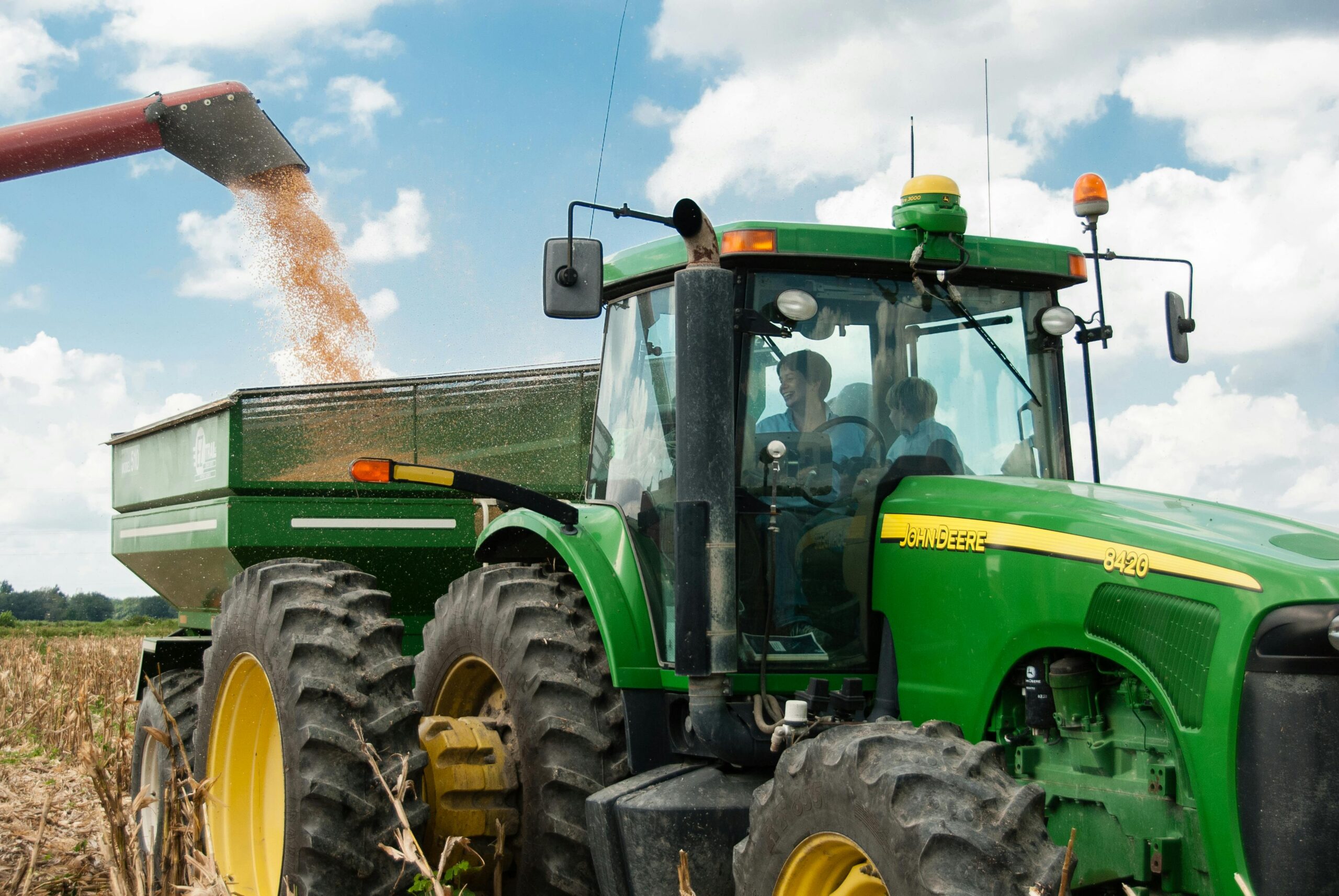 In the foreground, there is a John Deere green tractor with two people in the cab, to the left there is a green trailer that is collecting cut corn from a field. The sky is blue with lots of clouds