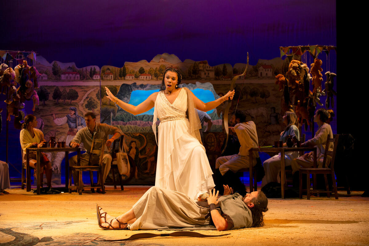 Stage setting with background singers in the shadows, the 2 main opera singers are lit up in the centre of the image with one person lying on the ground and the other standing over them