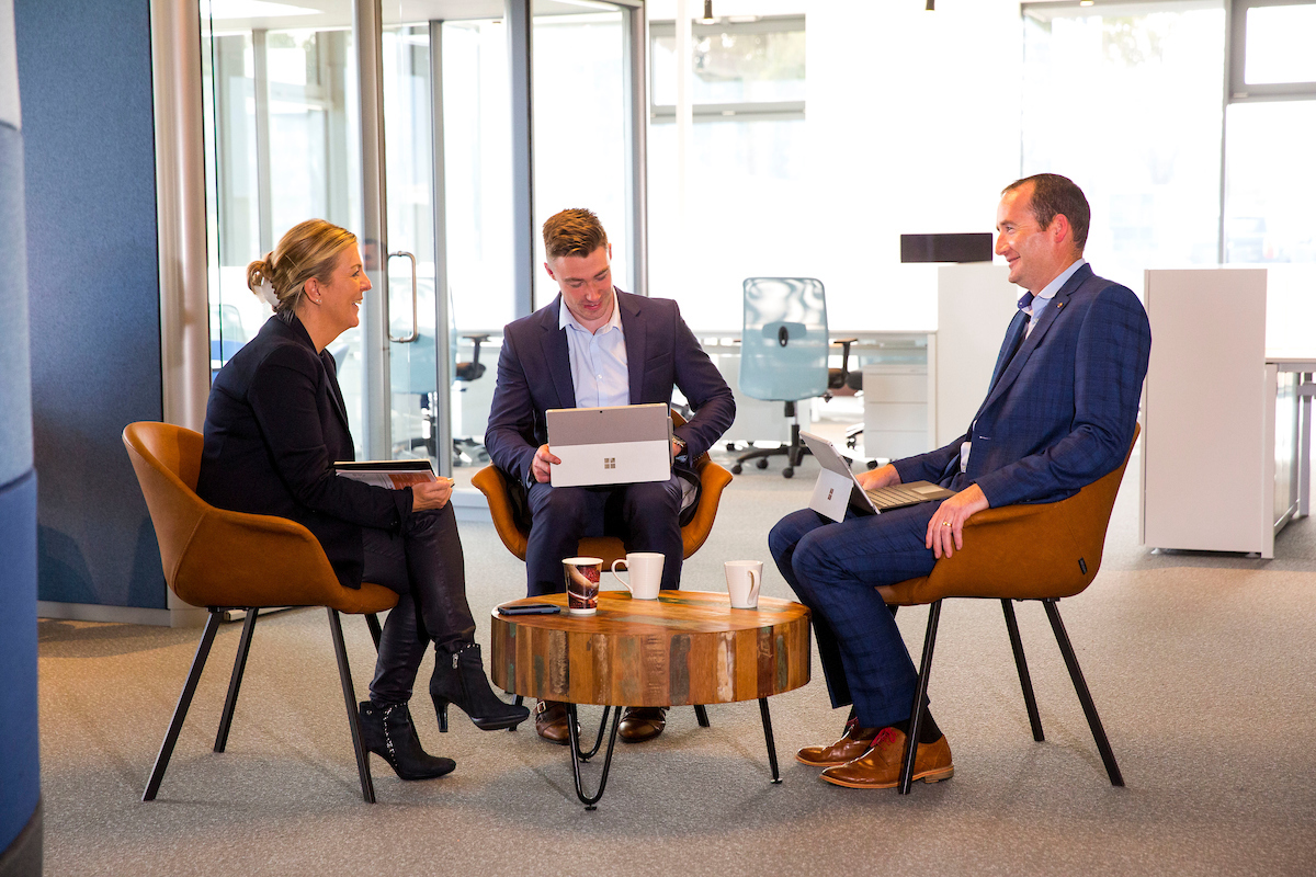3 business people gathered around a small round table. The man in the middle has a laptop on his lap, they are at the centre of the image