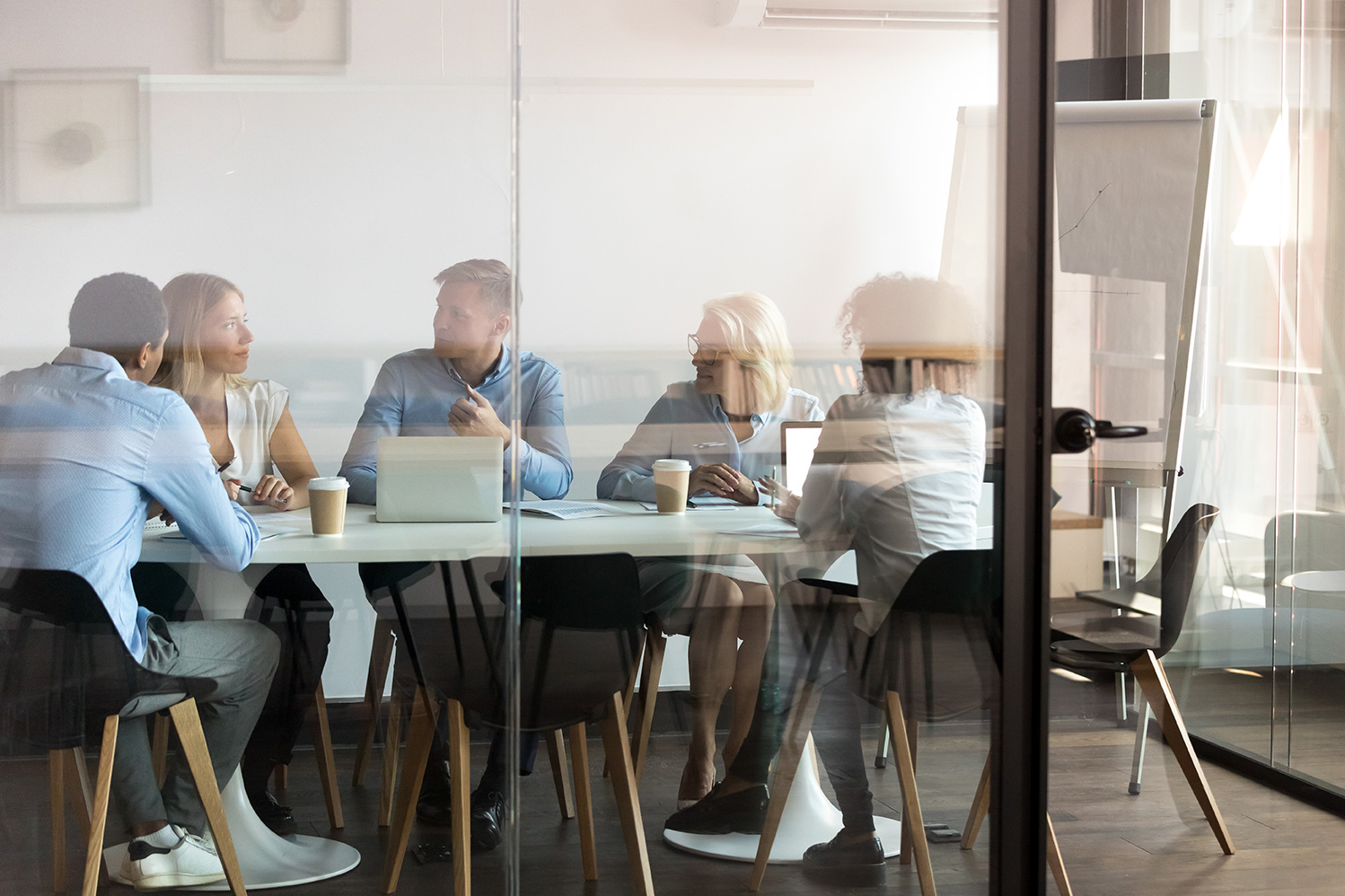 A modern boardroom with floor-to-ceiling glass windows/doors and a meeting is in session with multiple people sat around a long rectangular desk