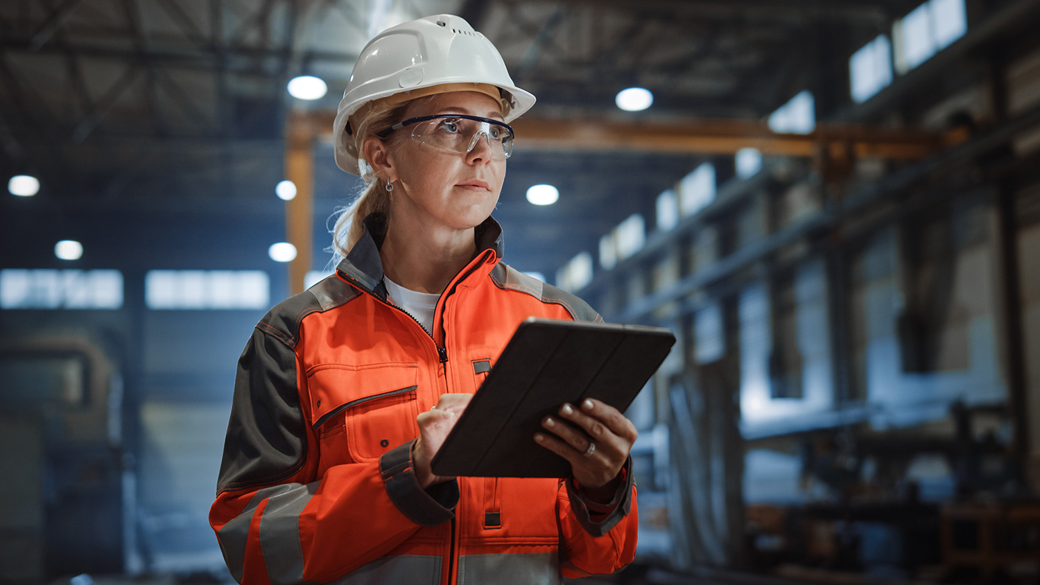 A lady wearing a hi-vis jacket, safety glasses and hard hat holds a tablet in a manufacturing warehouse