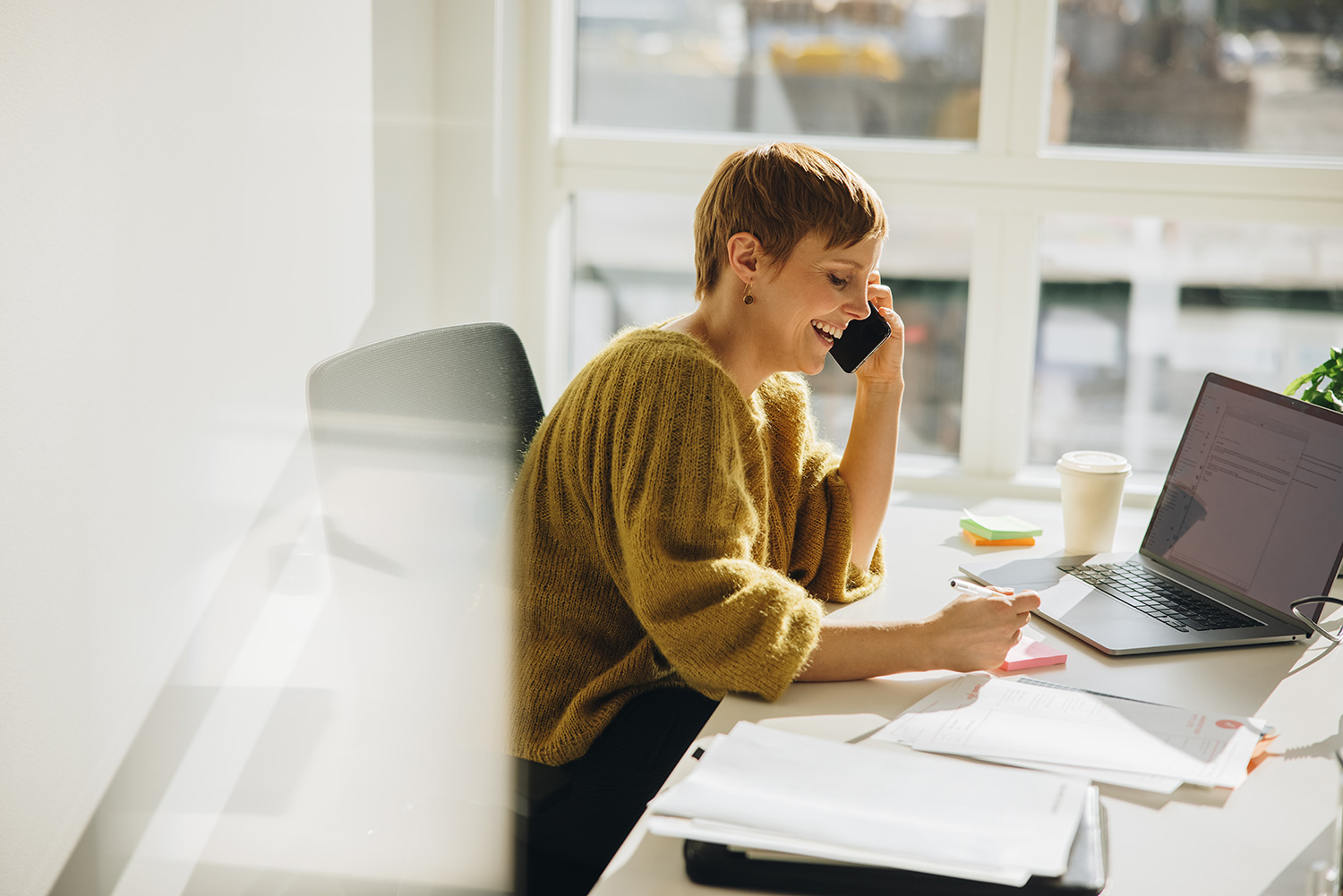 A woman sits at her office desk, laptop open and speaking on the phone. She is in a clinical, white office with large windows