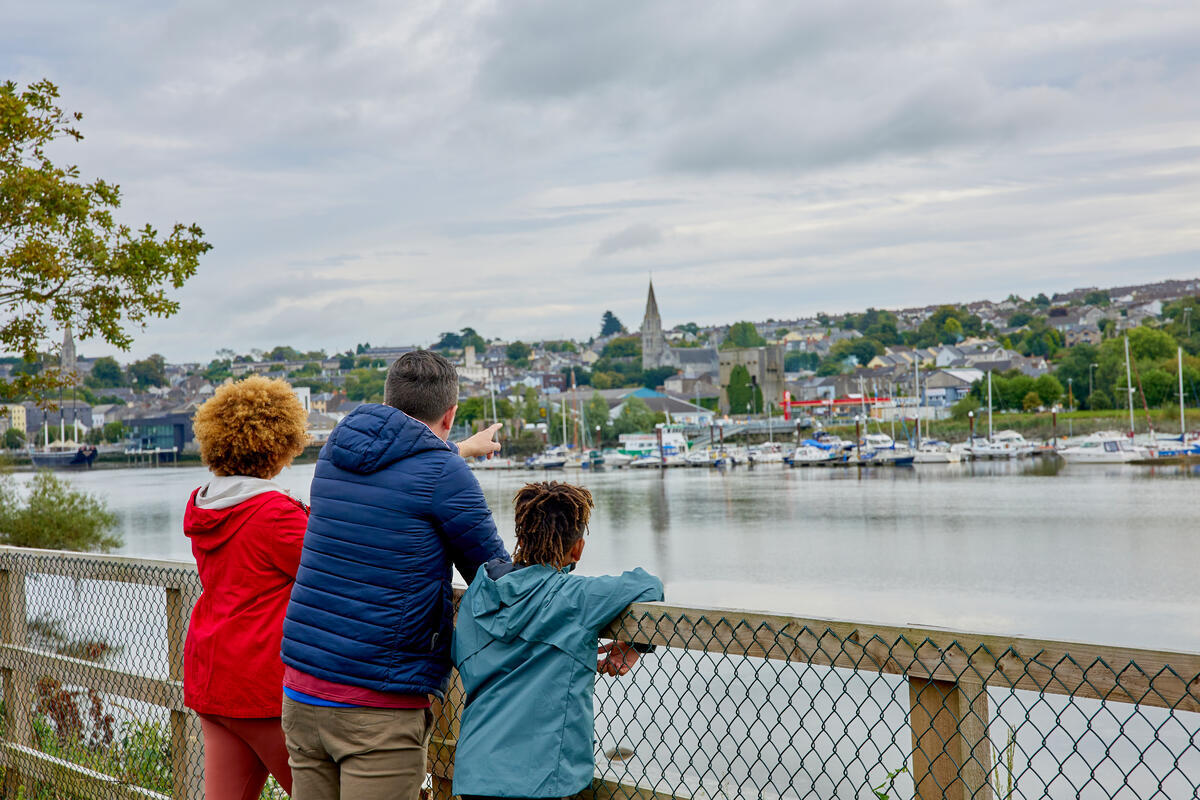 To the left foreground of the photo is a family of three, mother, father and young child. They are leaning on a fence overlooking the River Barrow in New Ross from the South East Greenway