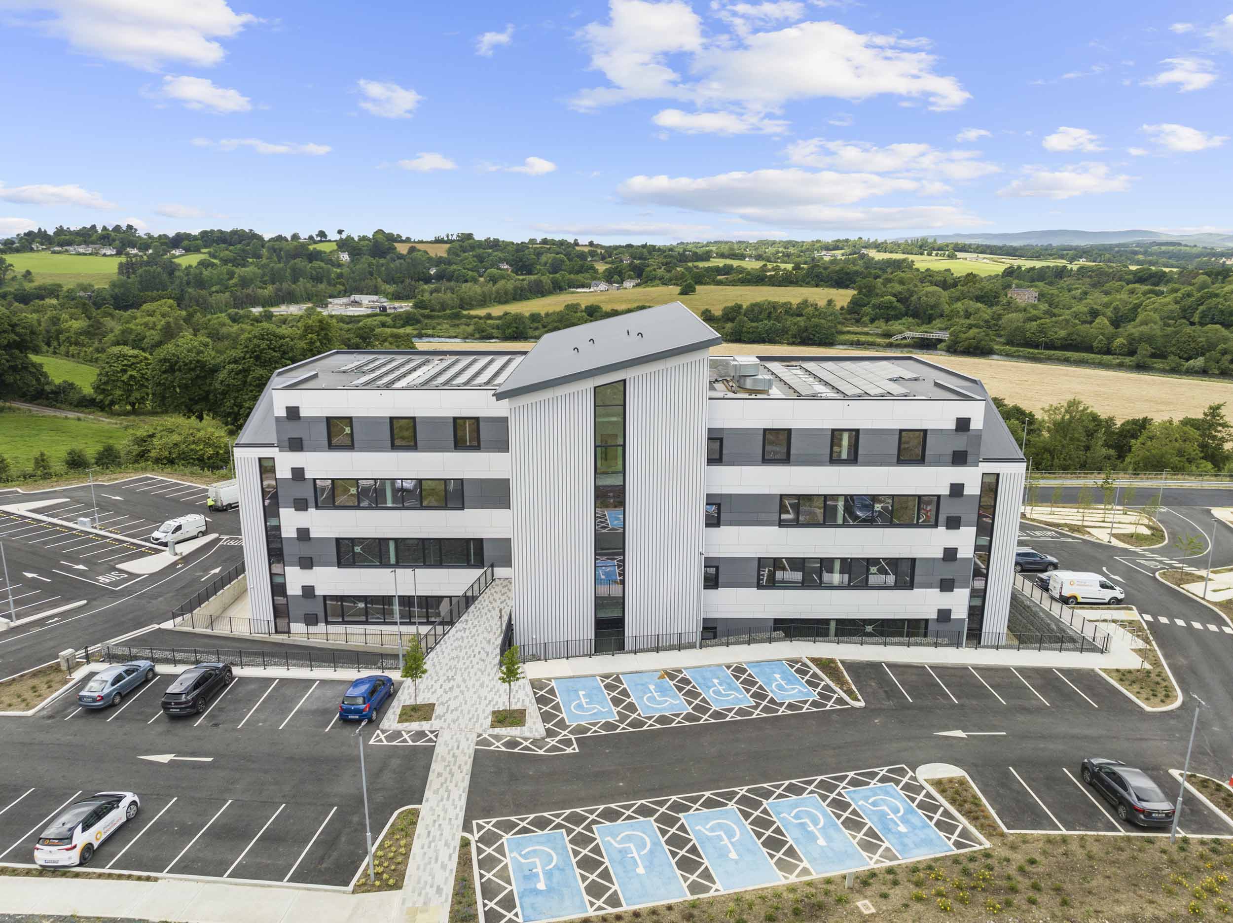 A large modern multi-storey building spans the centre of the image, the front façade of the building is a mixture of large glass panes and white panels. There are blue wheelchair parking spots to the foreground of the photo with another carpark to the left foreground.