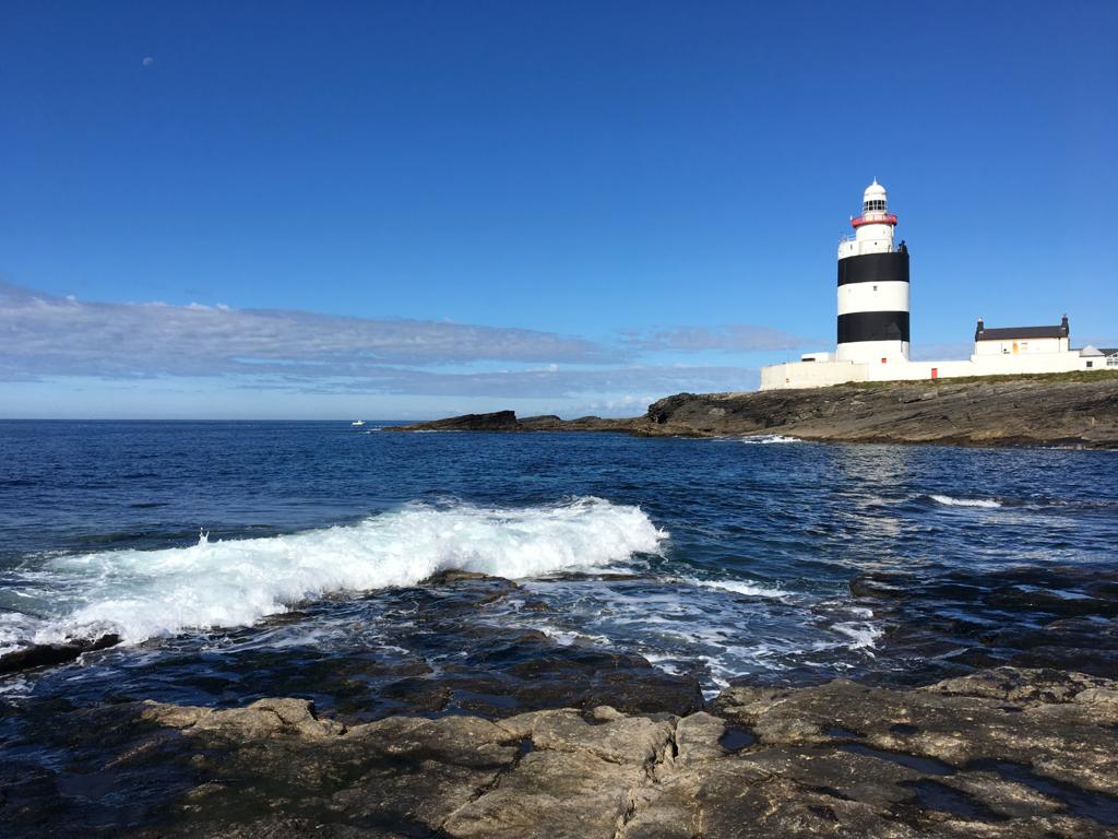 In the right of the image is Hook Lighthouse with its black and white horizontal stripes. It is a still, bright day on the peninsula with calm sea waters below and a blue sky above.