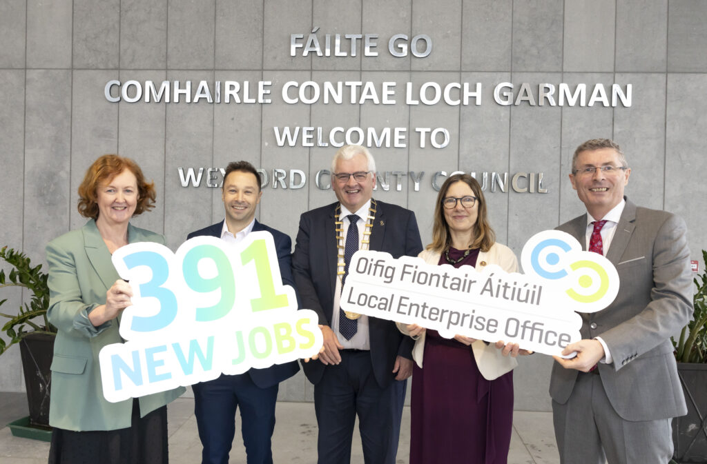 Local Enterprise Office team outside of the Wexford County Hall. Grey wall with mirrored lettering with 'Fáilte go Comhairle Contae Loch Garman' behind the 5 people