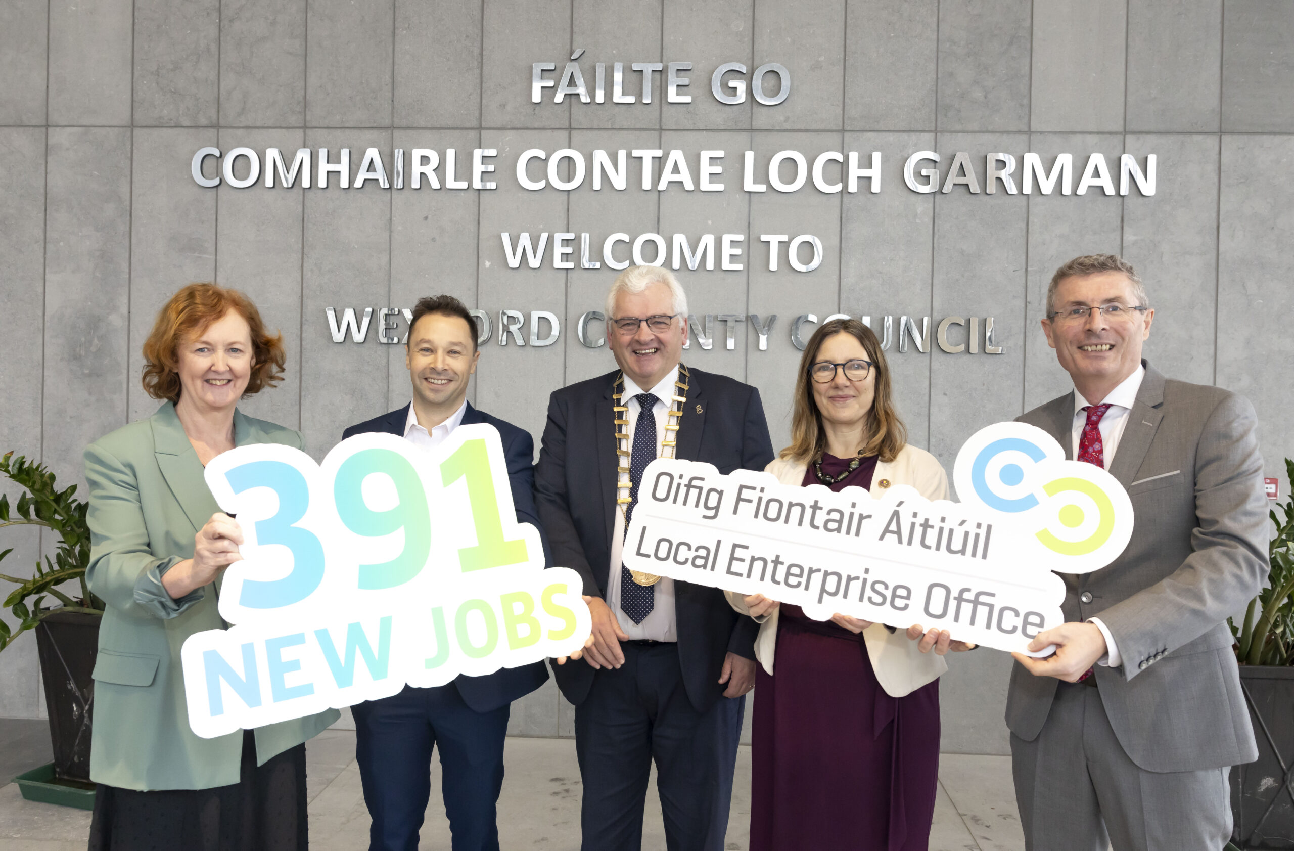 Local Enterprise Office team outside of the Wexford County Hall. Grey wall with mirrored lettering with 'Fáilte go Comhairle Contae Loch Garman' behind the 5 people
