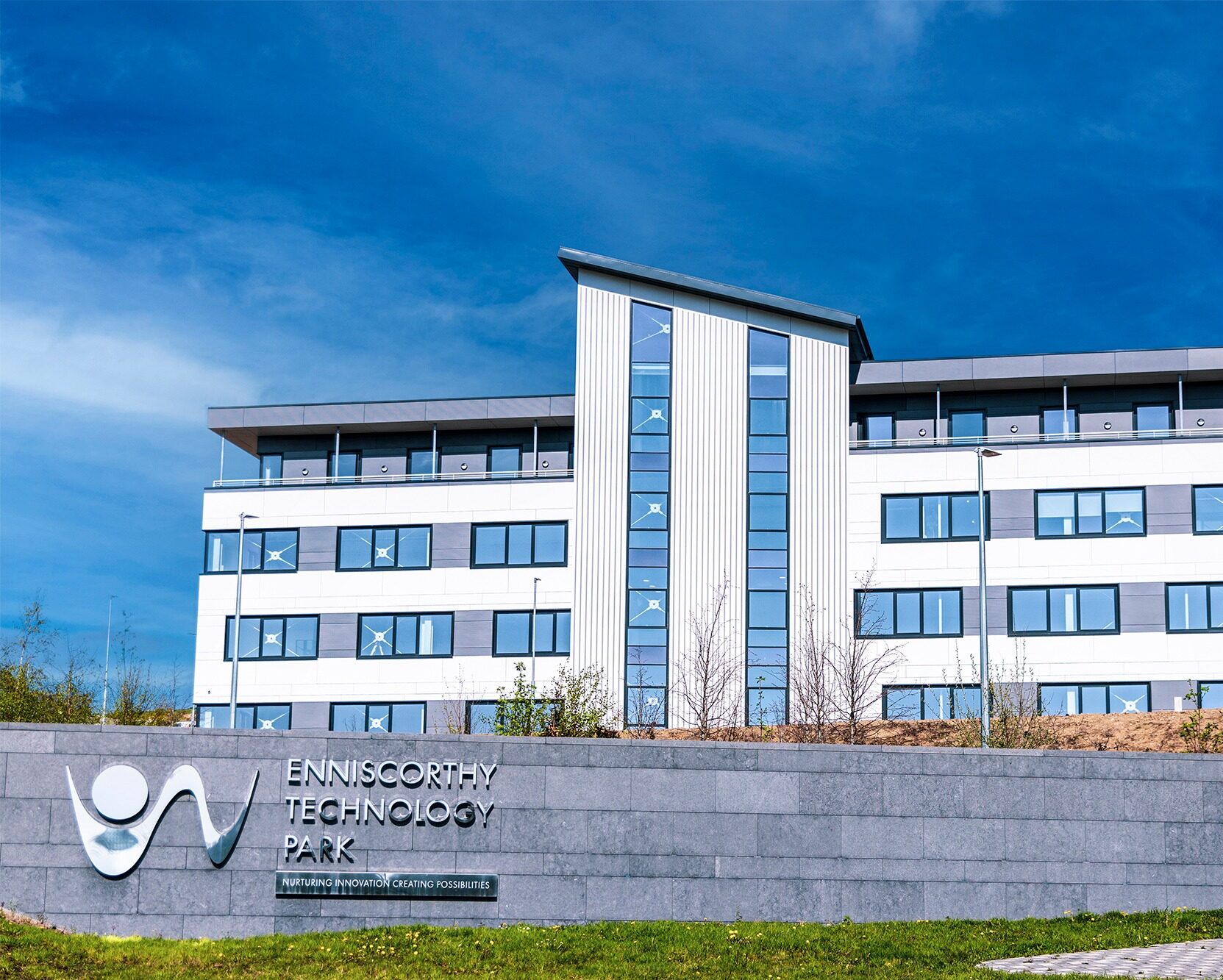 A large modern multi-storey building spans the centre of the image, the front façade of the building is a mixture of large glass panes and white panels. To the front foreground of the image is a grey, brick wall that has mirrored lettering showcasing the Wexford County Council logo and the name: Enniscorthy Technology Park.