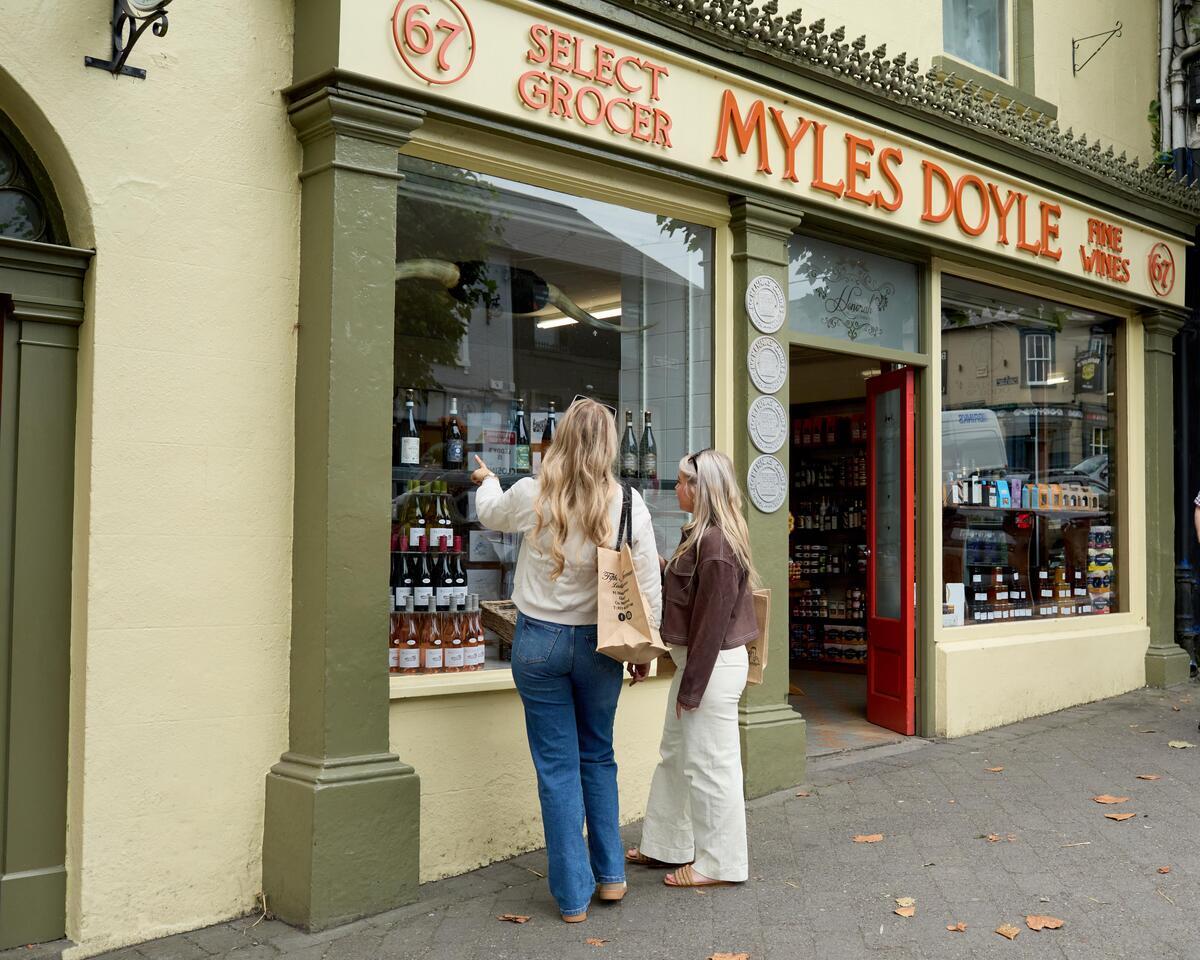 Two women look into a shopfront window to the left of the image. The shopfront is decorated with a vintage sign and reads Myles Doyle