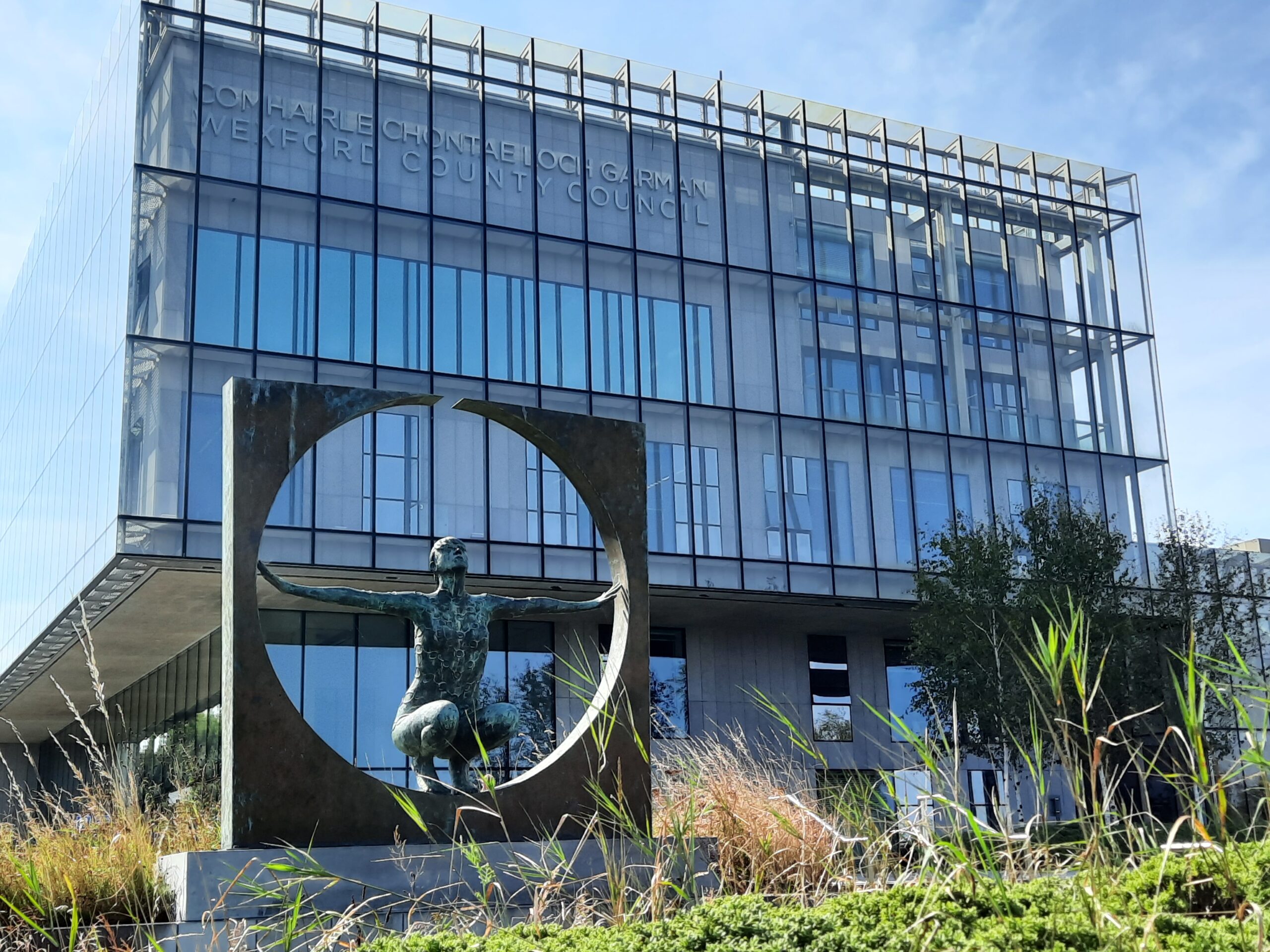 A front view of the Wexford County Council offices in the County Hall. The building is a multi-storey, glass fronted building with exposed metal rods as part of the façade. To the front of the building in the left foreground is a sculpture of a 3d square with a circle cut out in the centre. In the centre is a life-like sculpture of a lady with her arms out, holding both sides of the square. The piece is by Rowan Gillespie and is called 'Solstice'