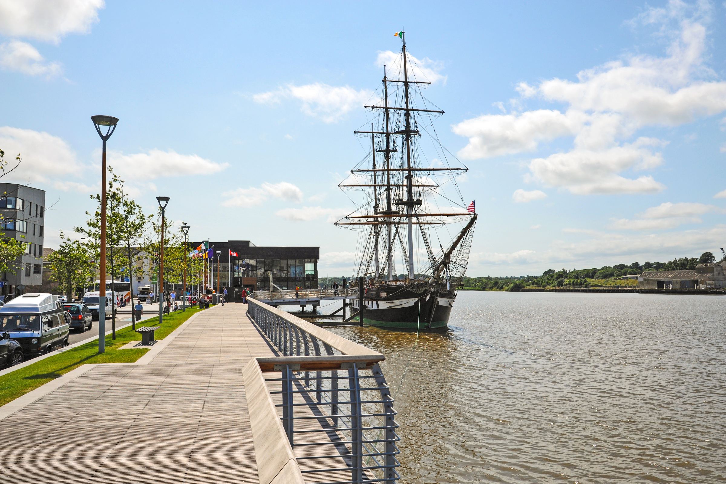 To the centre-right of the image is the Dunbrody Famine Ship, docked on the Barrow River which runs through New Ross town. To the left of the image is the boardwalk that leads into the Dunbrody Famine Ship Experience tourist centre. The sky is light blue with scattered clouds and it is a bright, sunny day.