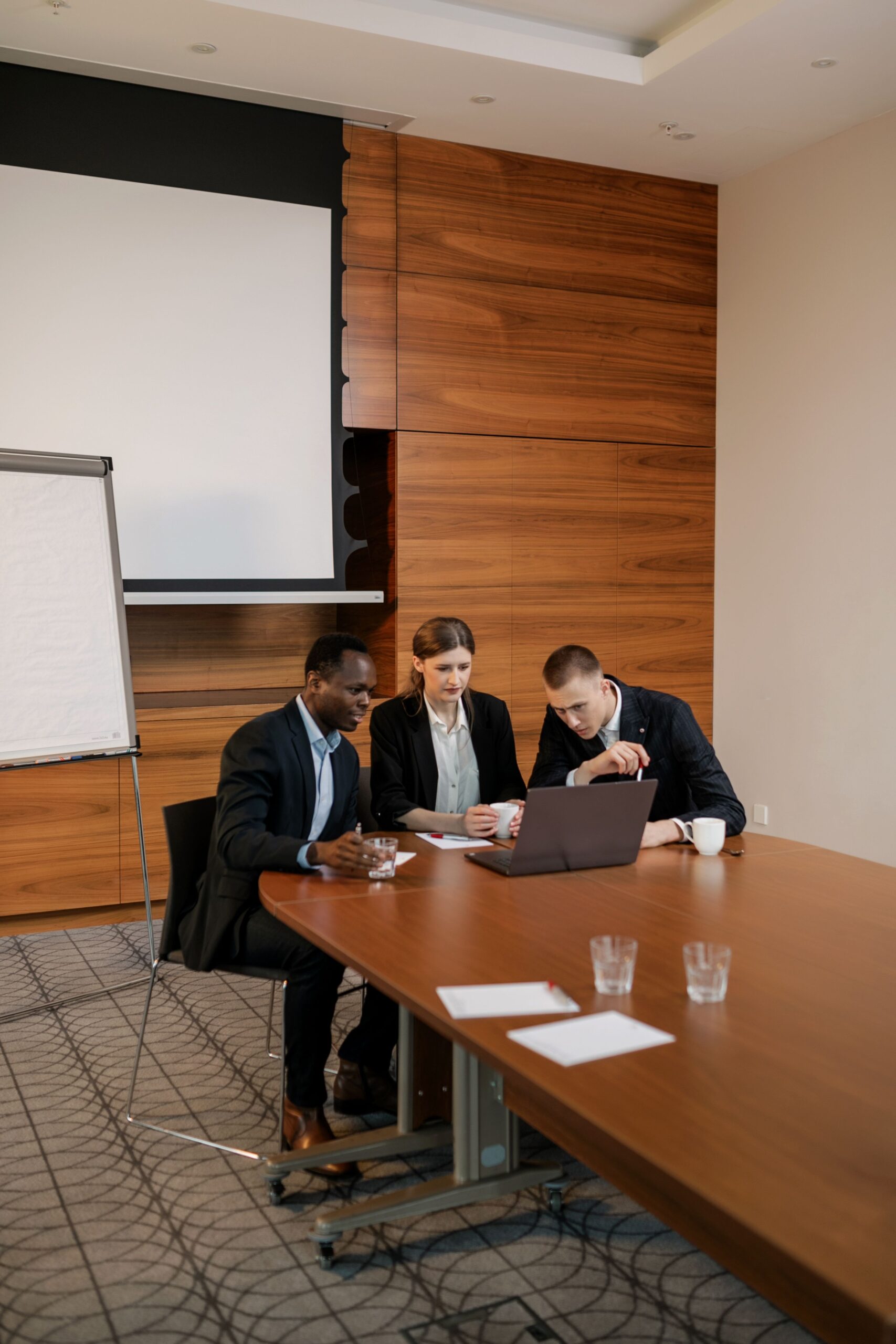 3 businesspeople sitting in front of a laptop in a professional boardroom setting. In deep discussion while examining the laptop