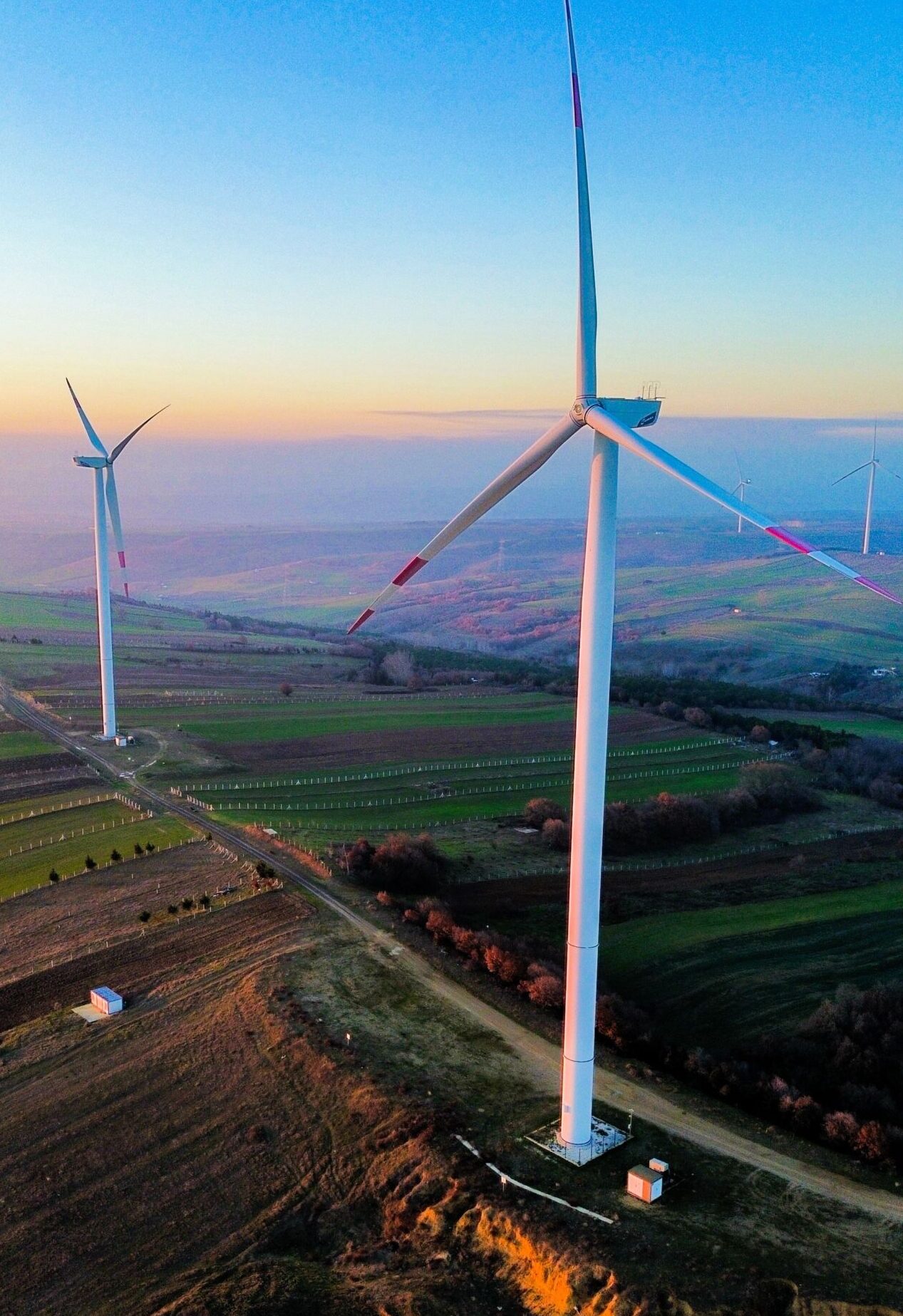 This is a drone shot from a wind farm. To the right foreground is one turbine with another one close by in the left background. They are set on grassland and the photo is taken at sunset. The sky is a light blue with a yellowy orange line across the centre of the image