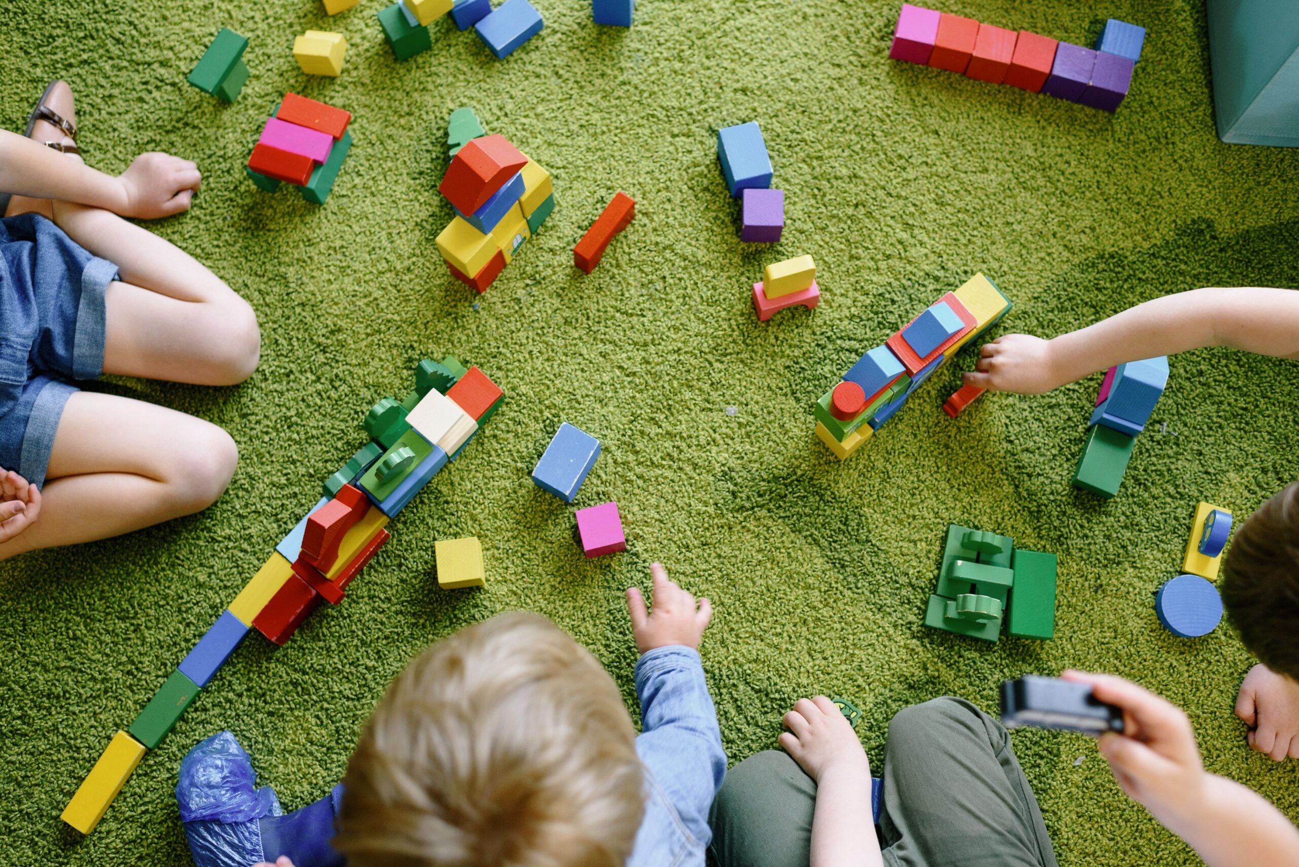 An overhead shot of a green carpet with lots of colourful children's building blocks. The tops of young children's heads are shown in the bottom centre of the overhead shot and there are children reaching their arms out to play to the right of the image