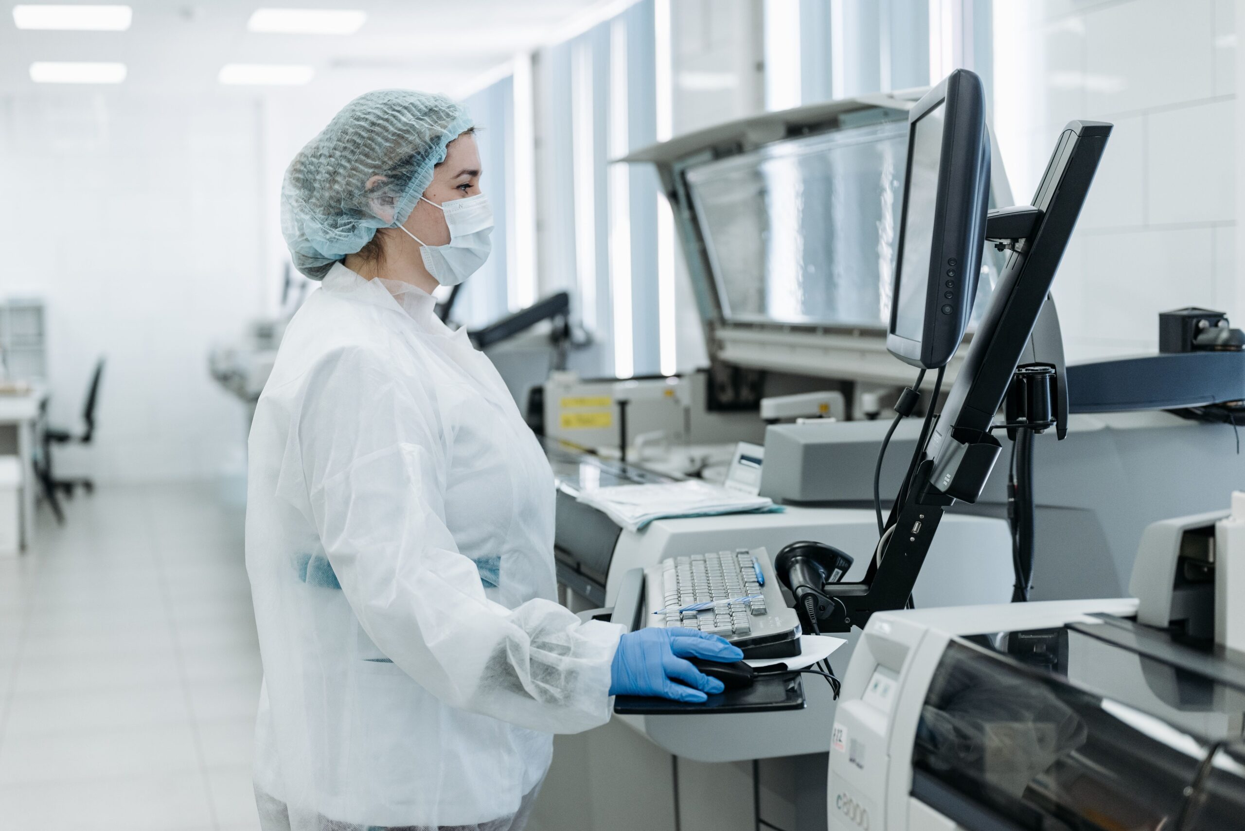a lab technician stands to the left of the image in a white room, they are gowned up with hairnet, surgical face mask and gloves, looking at the results on a computer attached to a large machine on the right-hand side of the image