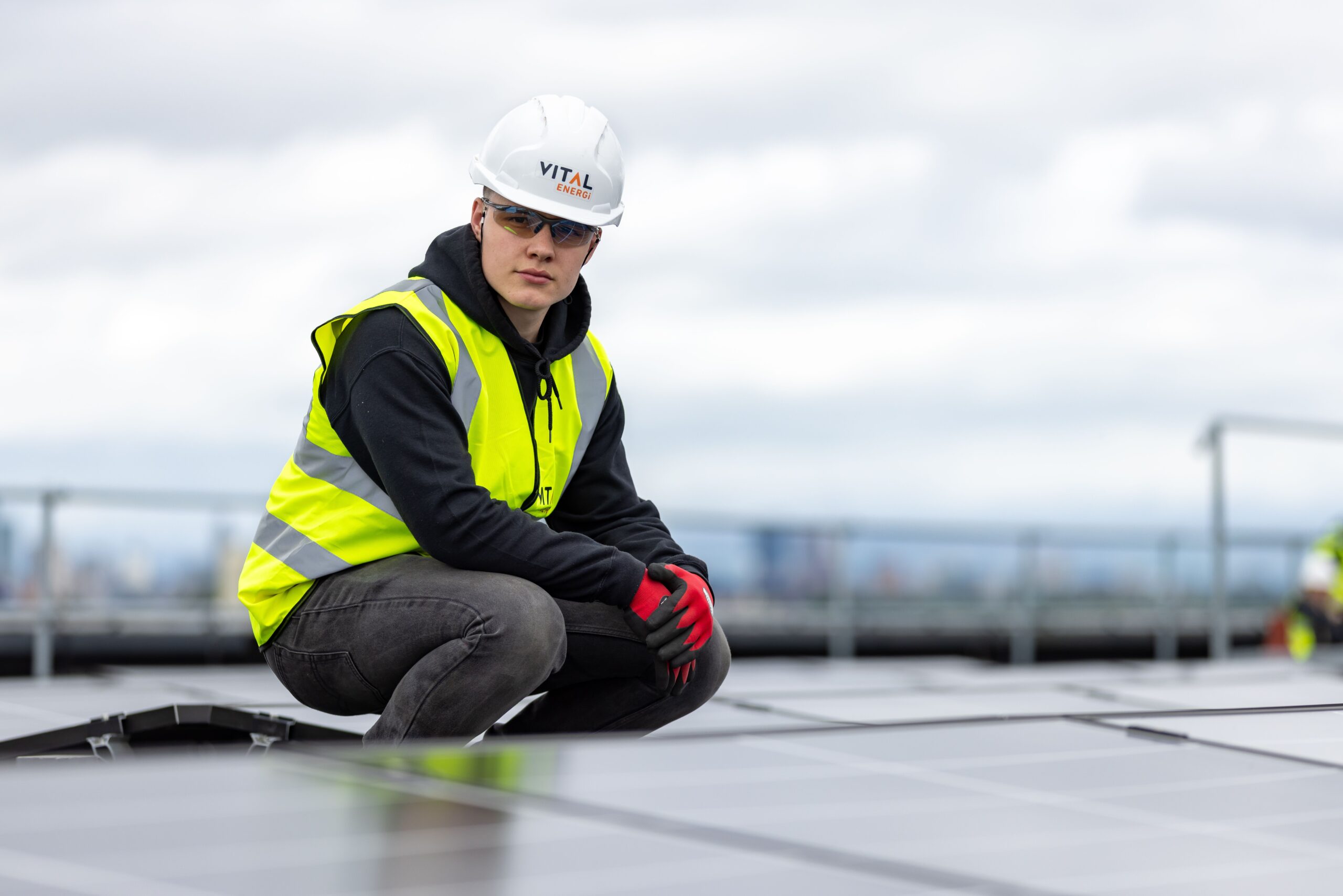 There is a solar power technician crouching down amongst a field of solar panels to the left of the photo. He is dressed in black clothes with a hi-vis vest and hard hat