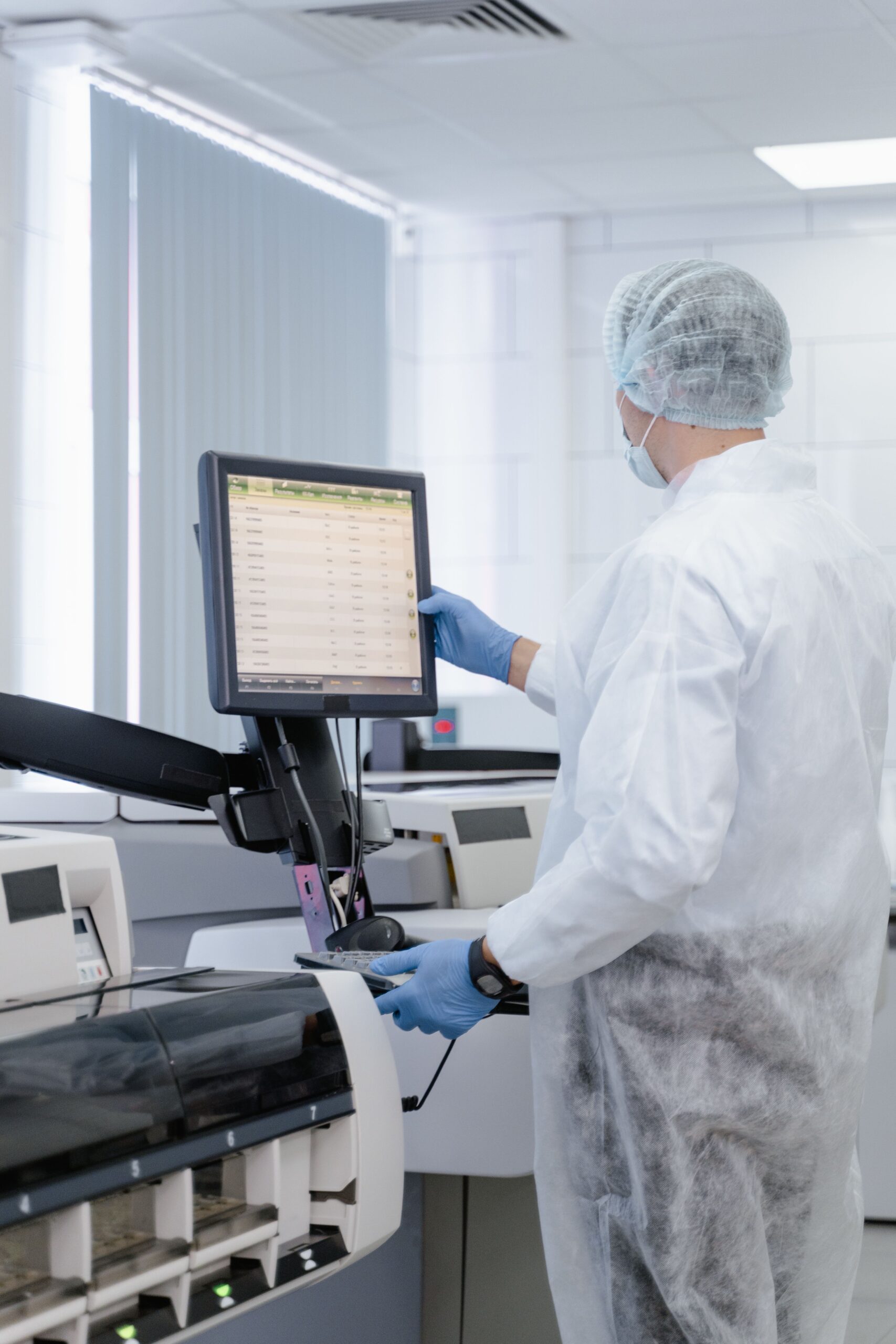 A man stands to the right of the image in full lab attire including medical gloves and hairnet. He is in a clinical white room beside a large machine to the left. He is looking at a computer screen analysing lab results
