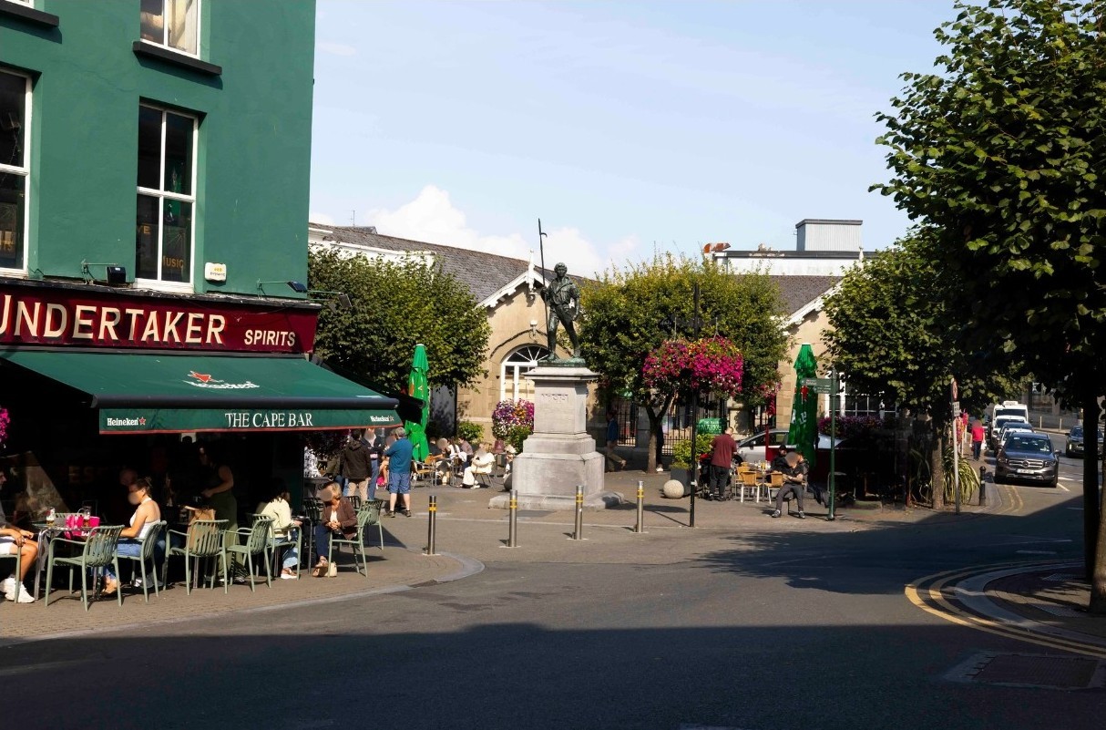 To the left is a pub with a canopy and outdoor seating, it is beside a main street and a Saturday market. There is a statue in the centre of the market square
