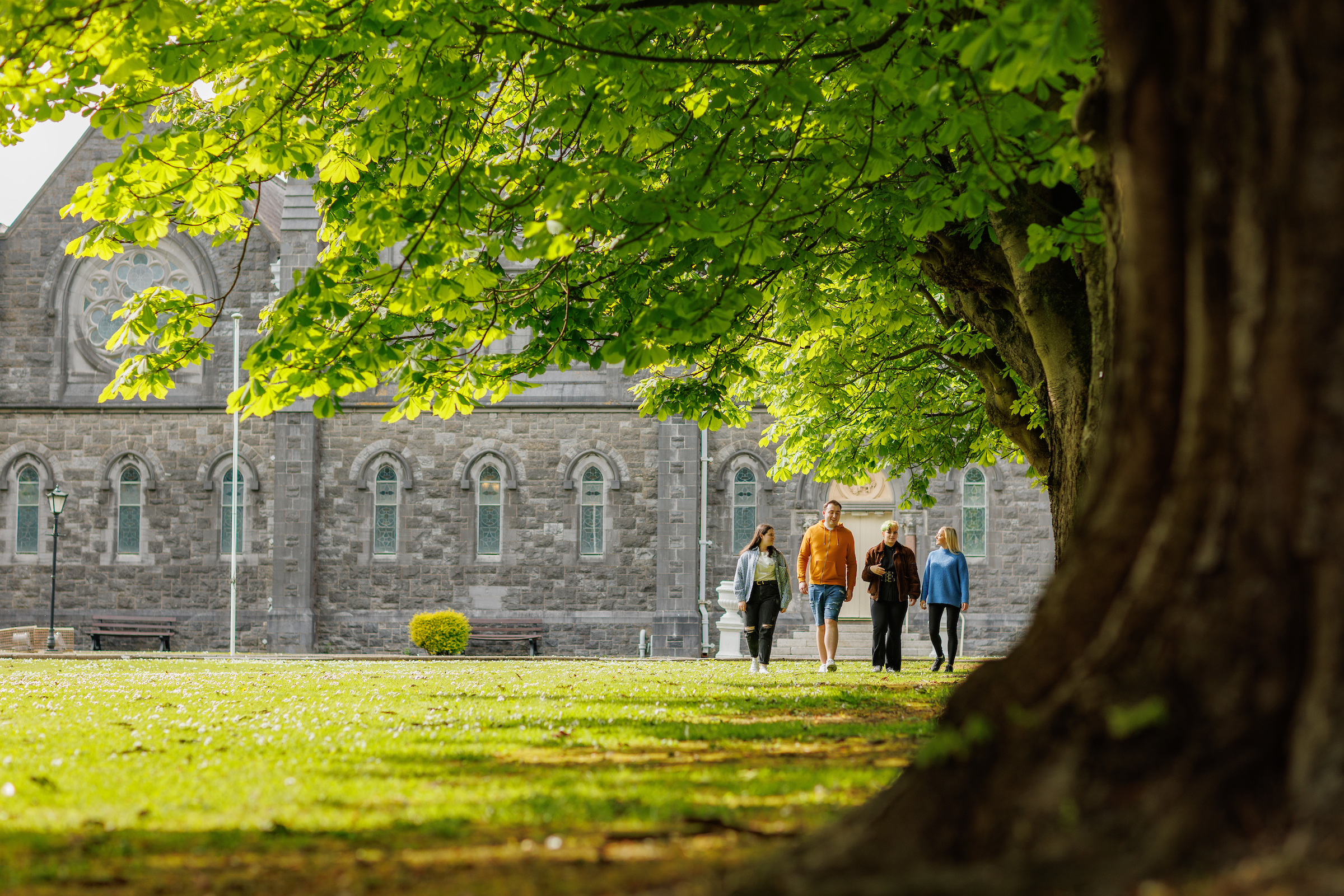 4 people walk facing the camera, away from the large 1800s building that houses SETU Wexford.