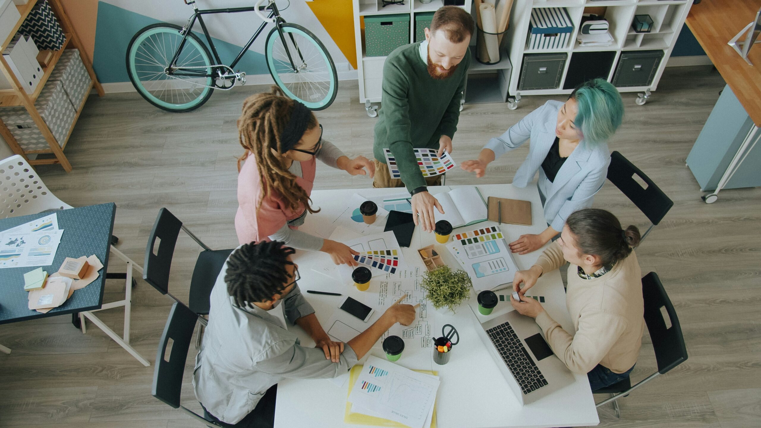 5 people crowded around a table, working on a project with laptops and phones