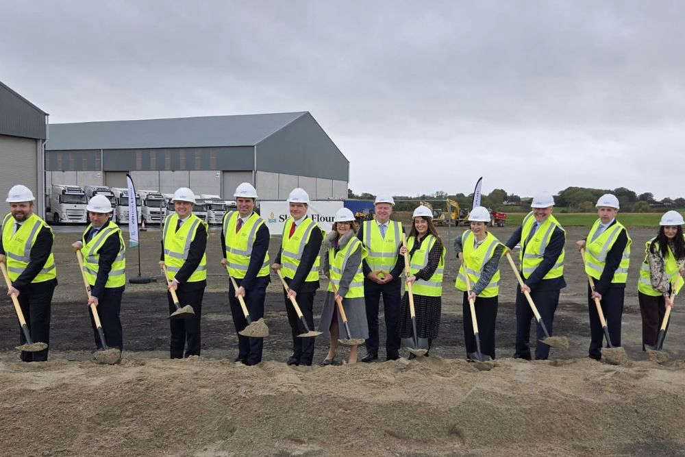 A group of people in hi-vis yellow vests and white hard hats standing in front of an industrial building turning the sod with shovels on a new development
